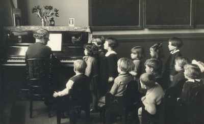 502155 Afbeelding van enkele kinderen van de Fröbelschool Ondiep 63 te Utrecht luisterend naar een leerkracht aan de piano.